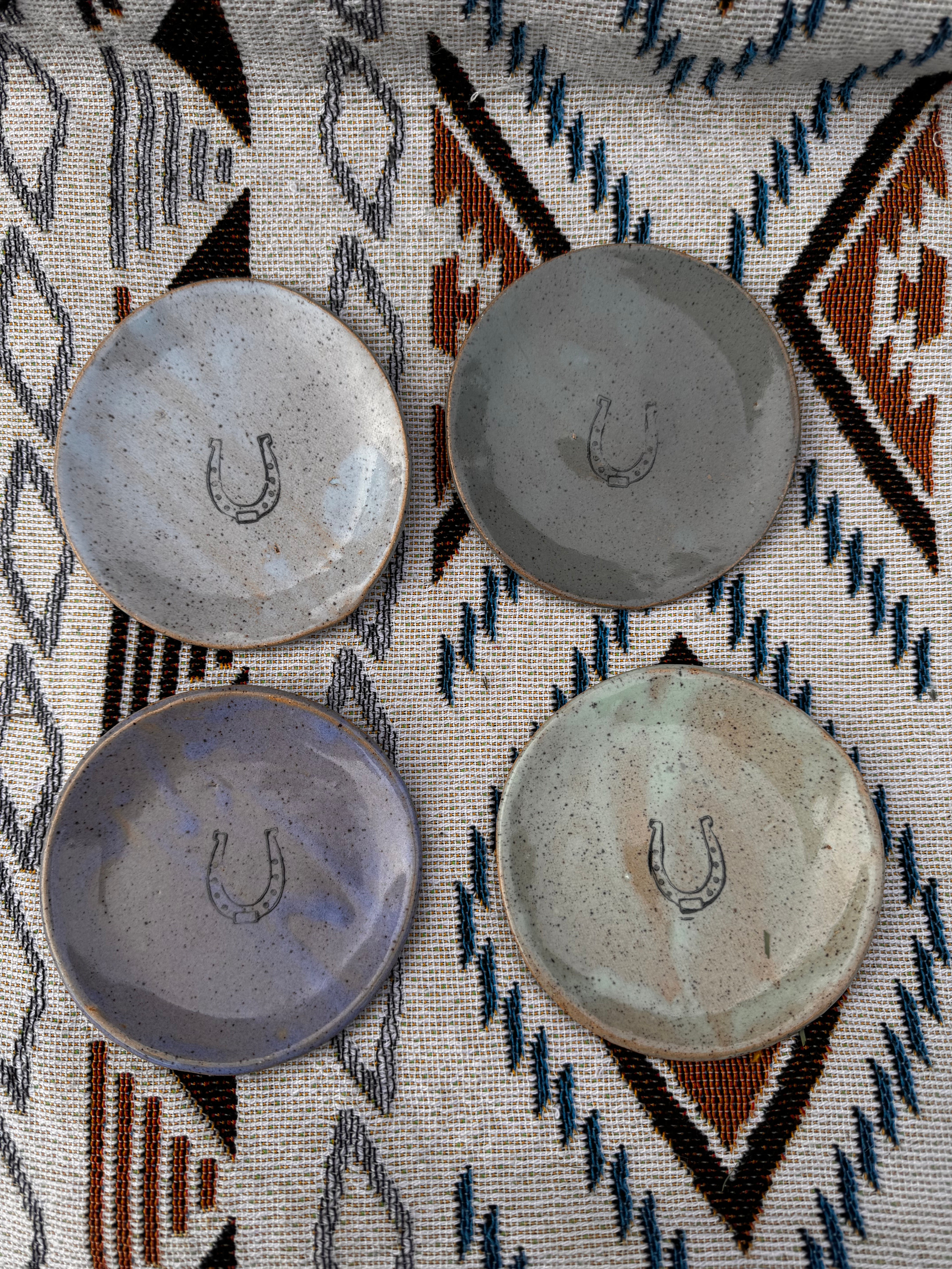 Handcrafted ring dish, about 3 inches in diameter, featuring a horseshoe design in the center, symbolizing good luck. Ideal for holding jewelry, tealights, dipping sauces, or jam on a cheese board, demonstrating its practicality and versatility. Each unique, artisan-made piece may vary slightly, showcasing the charm of handcrafted goods.