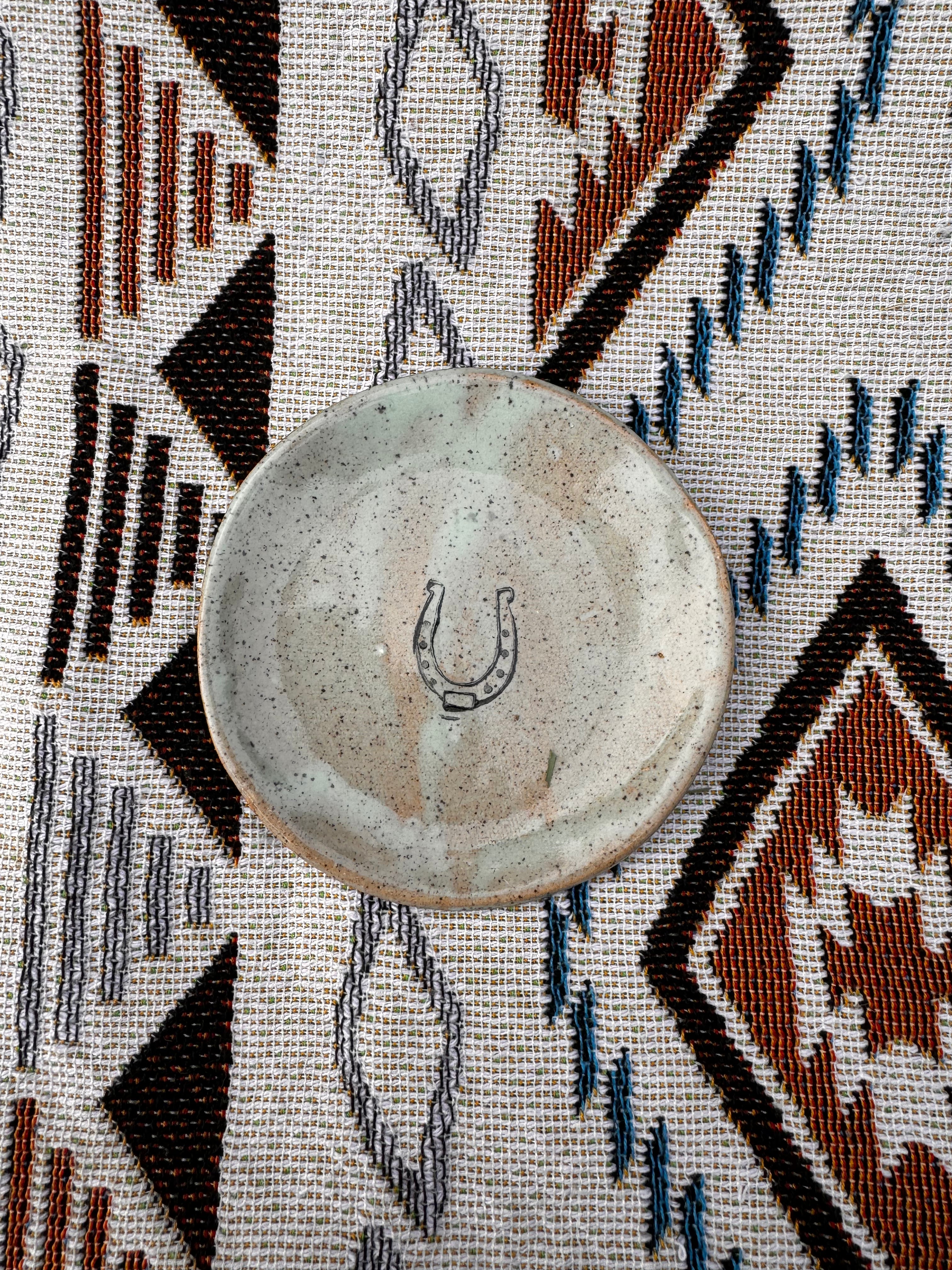 Handcrafted ring dish, about 3 inches in diameter, featuring a horseshoe design in the center, symbolizing good luck. Ideal for holding jewelry, tealights, dipping sauces, or jam on a cheese board, demonstrating its practicality and versatility. Each unique, artisan-made piece may vary slightly, showcasing the charm of handcrafted goods.