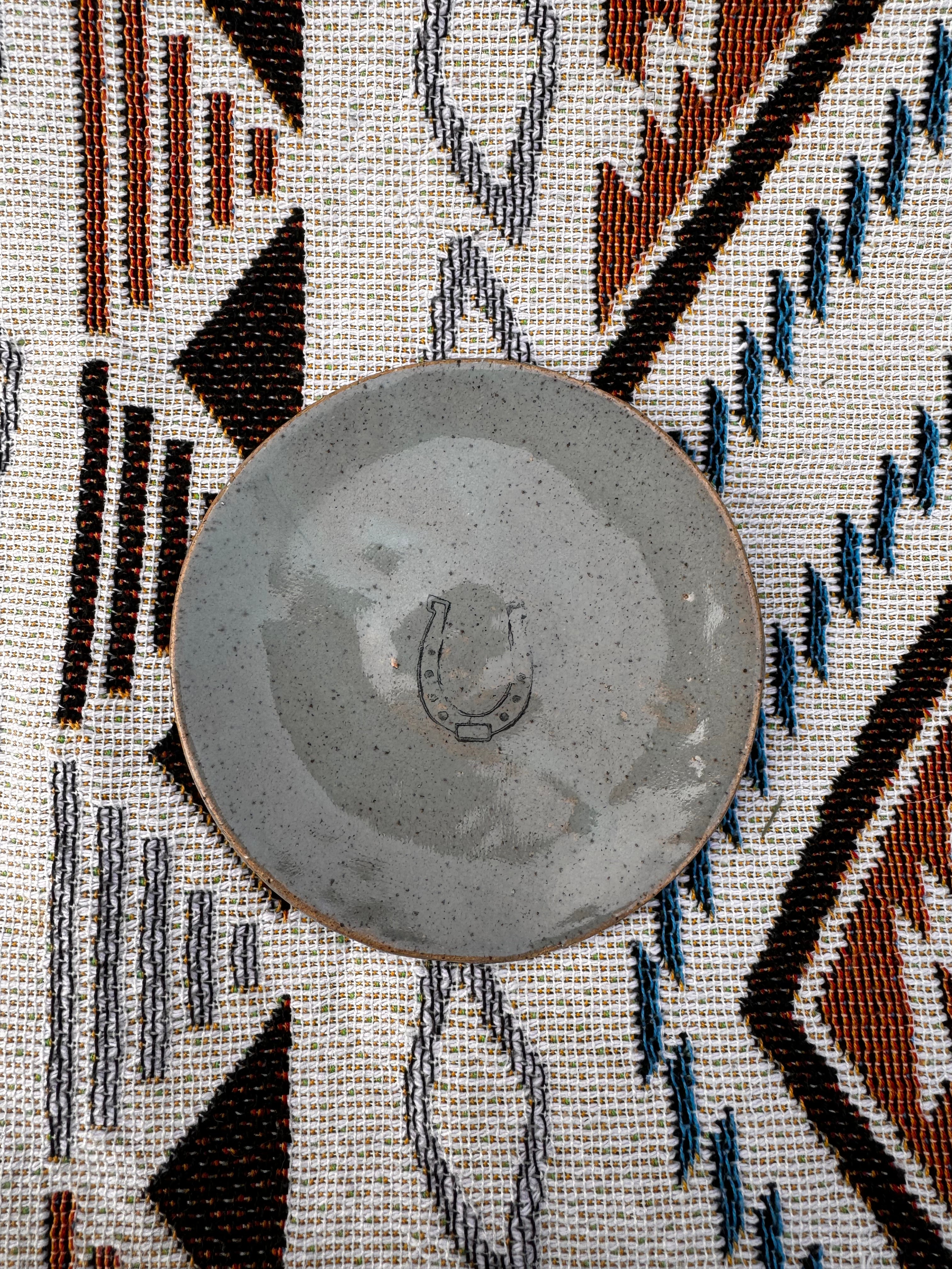 Handcrafted ring dish, about 3 inches in diameter, featuring a horseshoe design in the center, symbolizing good luck. Ideal for holding jewelry, tealights, dipping sauces, or jam on a cheese board, demonstrating its practicality and versatility. Each unique, artisan-made piece may vary slightly, showcasing the charm of handcrafted goods.