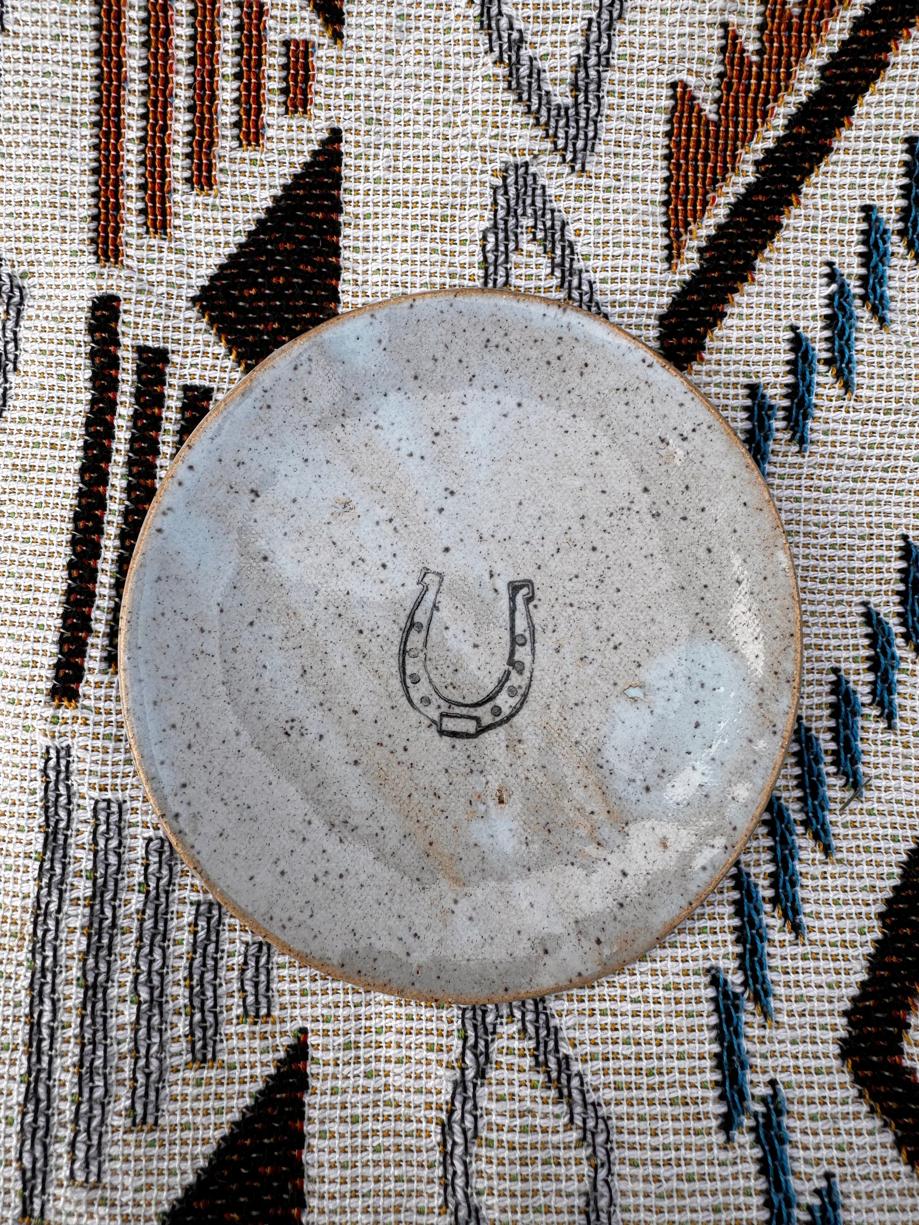 Handcrafted ring dish, about 3 inches in diameter, featuring a horseshoe design in the center, symbolizing good luck. Ideal for holding jewelry, tealights, dipping sauces, or jam on a cheese board, demonstrating its practicality and versatility. Each unique, artisan-made piece may vary slightly, showcasing the charm of handcrafted goods.